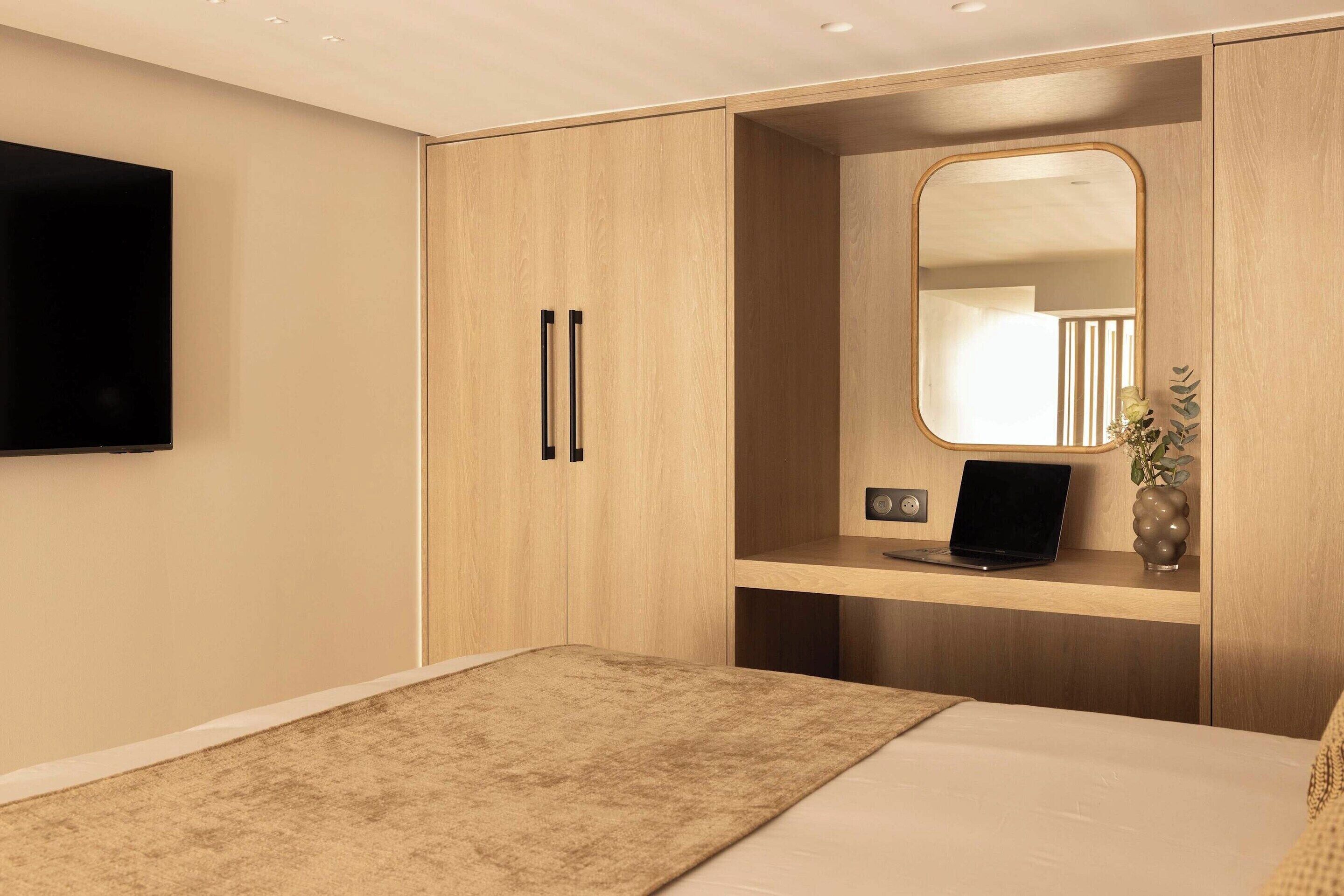 Een moderne slaapkamer met een bed, een aan de muur bevestigde televisie, een houten garderobe, een spiegel en een laptop op een plank.