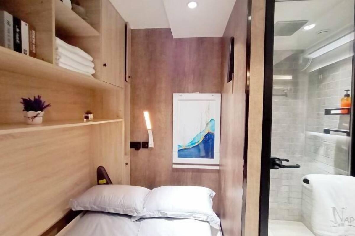 A compact hotel room with a bed, a small shelf with books and a potted plant, a painting on the wall, and a glass shower area.