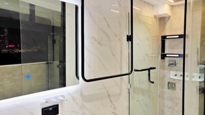 A modern bathroom with a glass shower enclosure, a marble wall, and a window overlooking a cityscape.