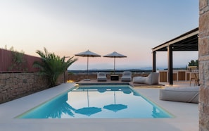 A pool with a clear view of the mountains, outdoor seating with umbrellas, and a stone wall.