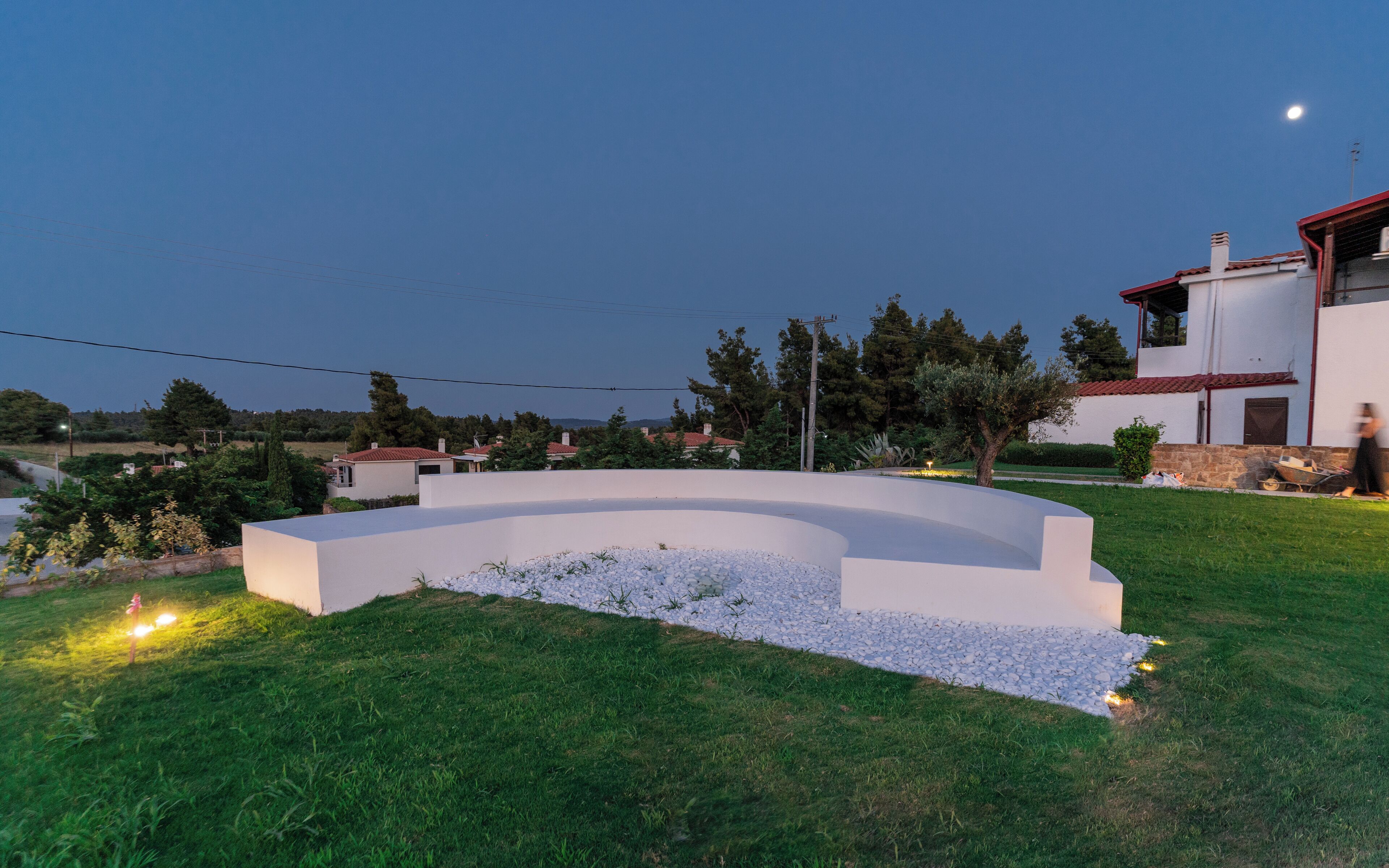 A modern outdoor seating area with a curved white structure, surrounded by a grassy lawn and a stone border.
