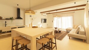 A modern kitchen with a central island, wooden chairs, and a dining table. There is a living area with a sofa and a TV.