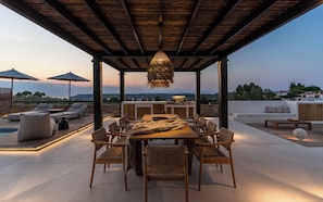 An outdoor patio with a wooden table and chairs, a hanging light fixture, and a view of the landscape.