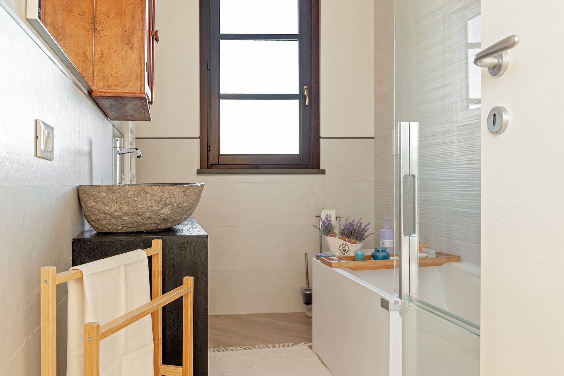 A compact bathroom with a wooden cabinet, a stone basin, a towel rack, a glass shower enclosure, and a small shelf with toiletries.
