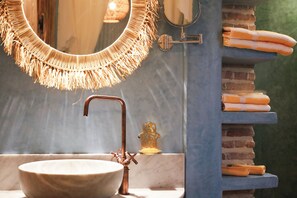 A bathroom with a round mirror, a copper faucet, a bowl sink, and a stack of towels on a shelf. - Riad 10 Mandarin (Marrakech)