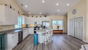 A spacious kitchen with white cabinetry, a central island with bar stools, a stainless steel dishwasher, and a large window providing a view of the outdoors.