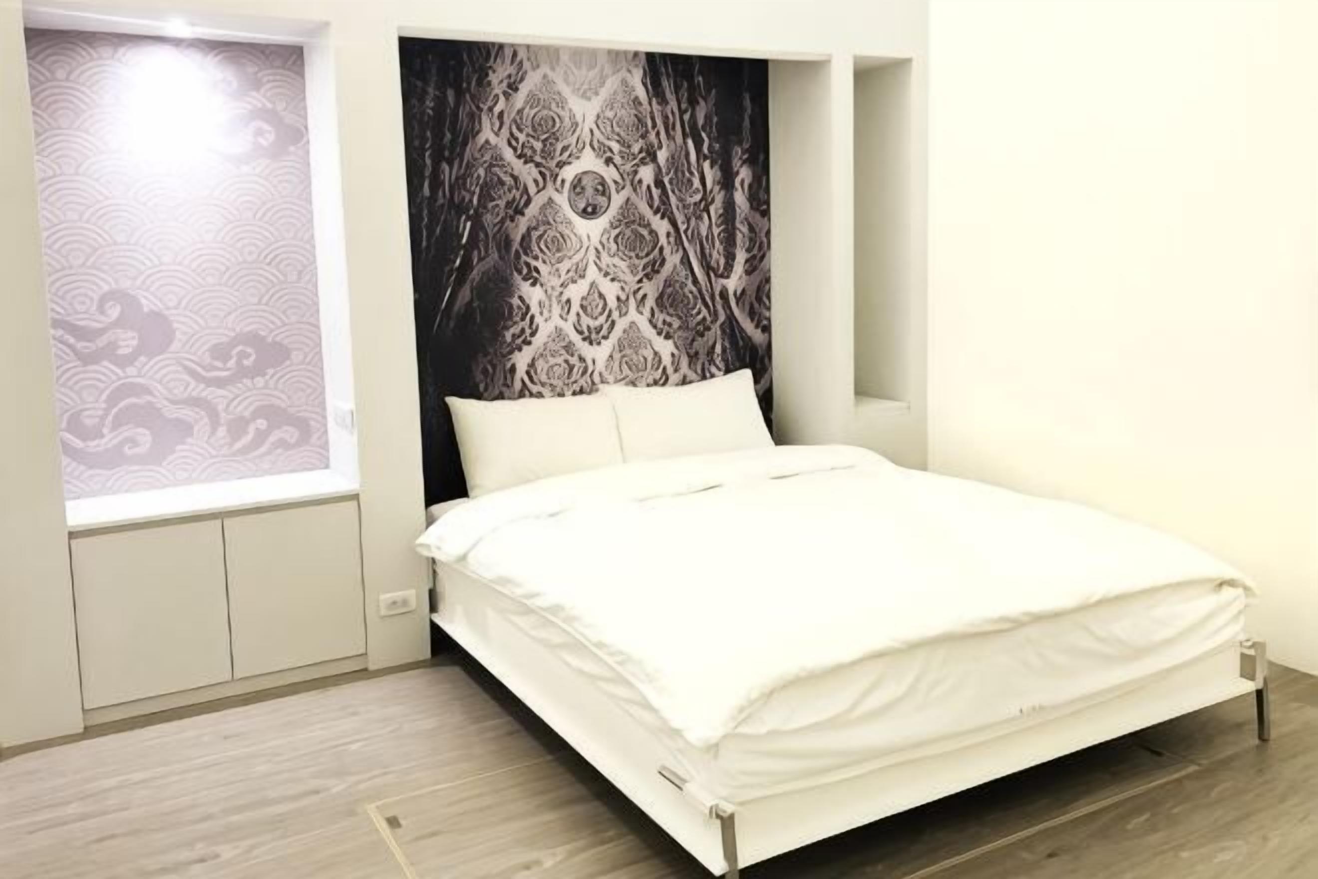 A bedroom with a bed, a window with patterned curtains, and a wall with a large abstract design.