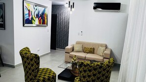 Una sala moderna con un sofá color crema, dos sillones estampados, una mesa de centro de vidrio y un cuadro abstracto y colorido en la pared.
