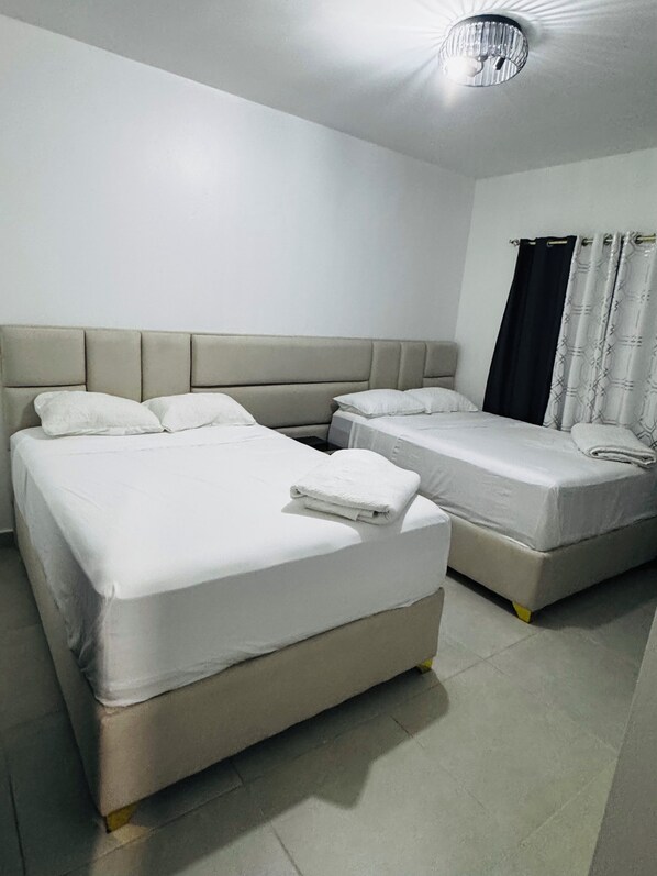Ein Hotelzimmer mit zwei Betten, einem Kopfteil, einer Deckenleuchte und einem Fenster mit Vorhängen.