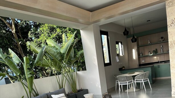 Un espacio moderno para estar con un sofá gris, una mesa de centro y una mesa de comedor con sillas. Se observa un ventilador de techo, una planta en maceta y una cocina al fondo.