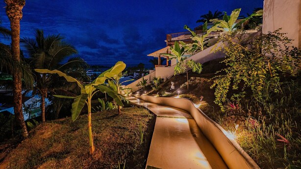 A nighttime view of a hillside with a water slide, palm trees, and a building with a red roof.