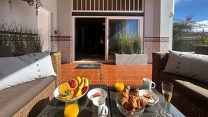 Una terraza con muebles de mimbre, una mesa de desayuno con frutas, croissants y bebidas, y una vista del interior a través de una puerta de vidrio.
