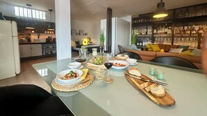 Una cocina moderna con comedor, que incluye una mesa de vidrio con comida y bebidas, una barra con botellas de licor y una zona de estar acogedora con cojines.