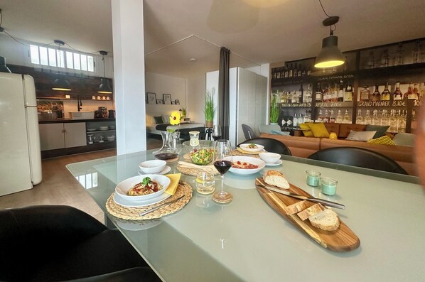 A modern kitchen with a dining area, featuring a glass-top table with food and drinks, a bar area with liquor bottles, and a cozy seating area with cushions.