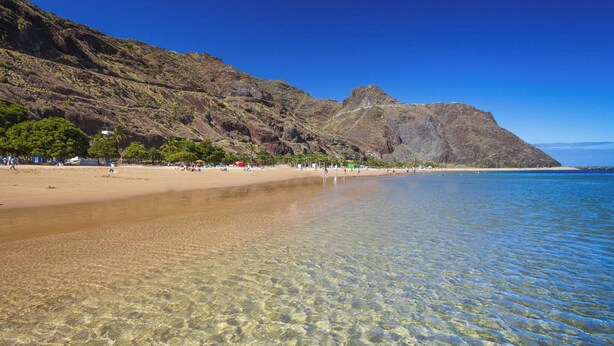 Una playa con aguas claras y poco profundas, una orilla arenosa y montañas al fondo.