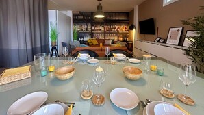 Un comedor con una mesa de vidrio dispuesta para seis personas, rodeado por un sofá y una estantería llena de libros y botellas.