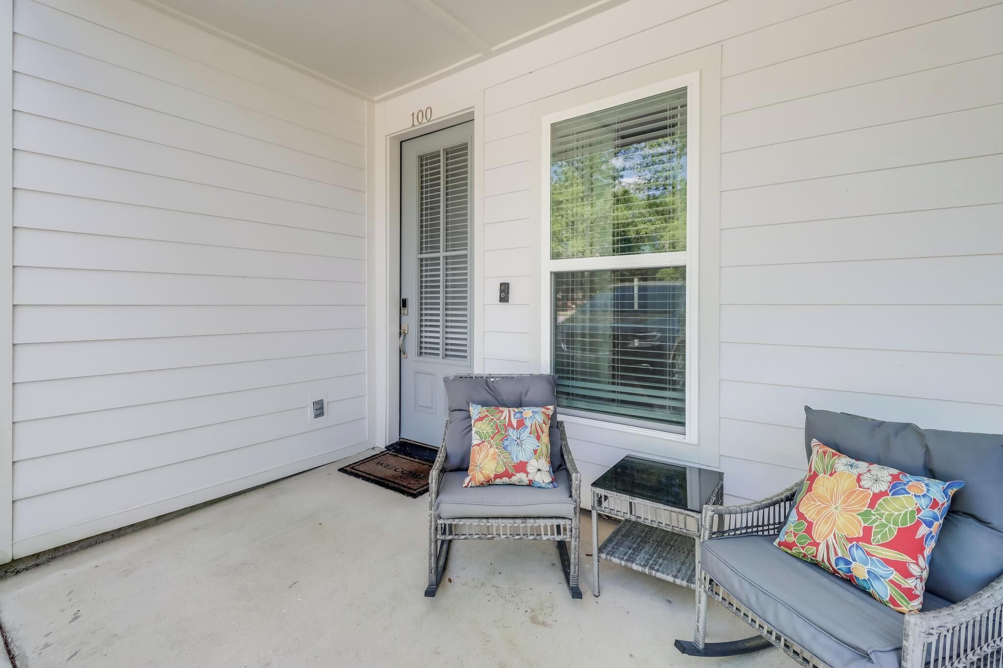 A porch with two wicker chairs, one with a colorful cushion and another with a dark cushion, and a small table.