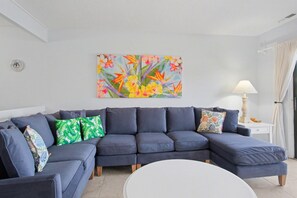 A living room with a dark blue sofa, a white coffee table, and a colorful floral painting on the wall. - 1611 Port Villa @ South Beach - Waterfront Villa (Hilton Head Island)