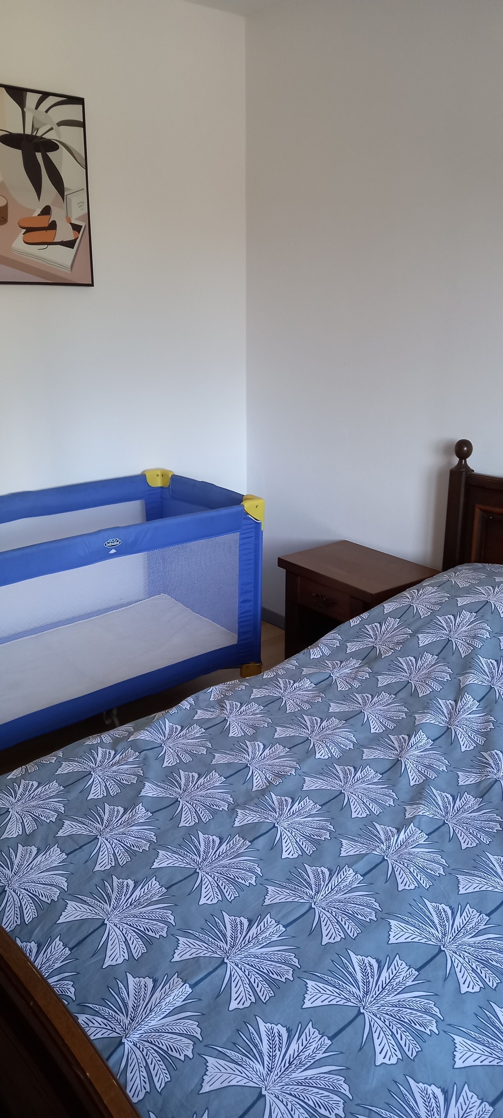 Un dormitorio con una cama, una colcha de edredón azul y blanco, una cama nido azul y blanca, una mesita de noche de madera y una obra de arte en la pared.