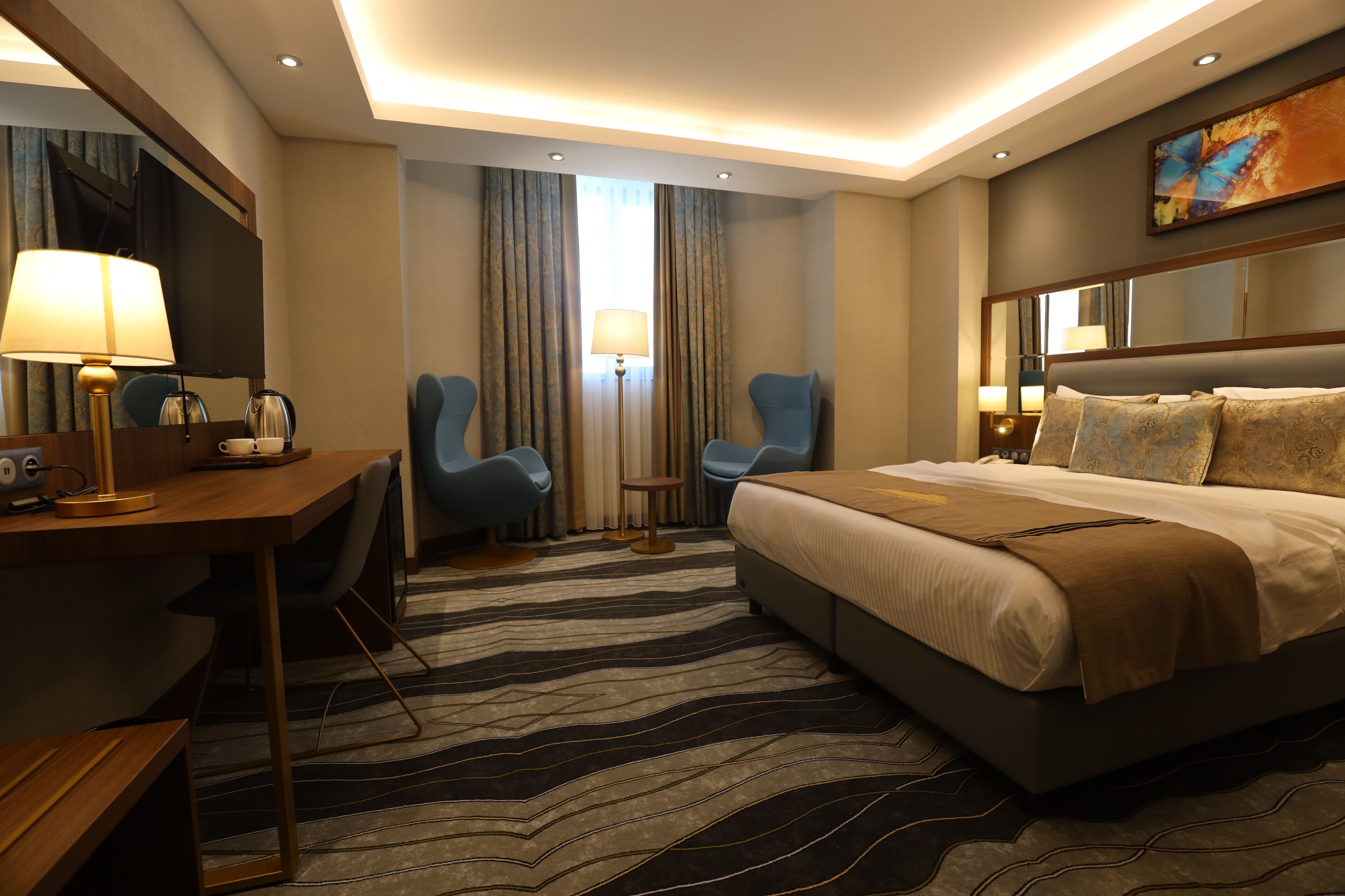 A hotel room with a large bed, two armchairs, a desk with a lamp, and a large mirror.