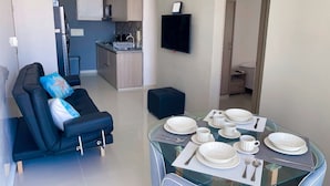 A modern living room with a glass dining table set for four, a black sofa with cushions, and a kitchen in the background.
