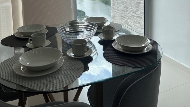 A dining area with a glass table, black chairs, and a view of a river and buildings through a large window.