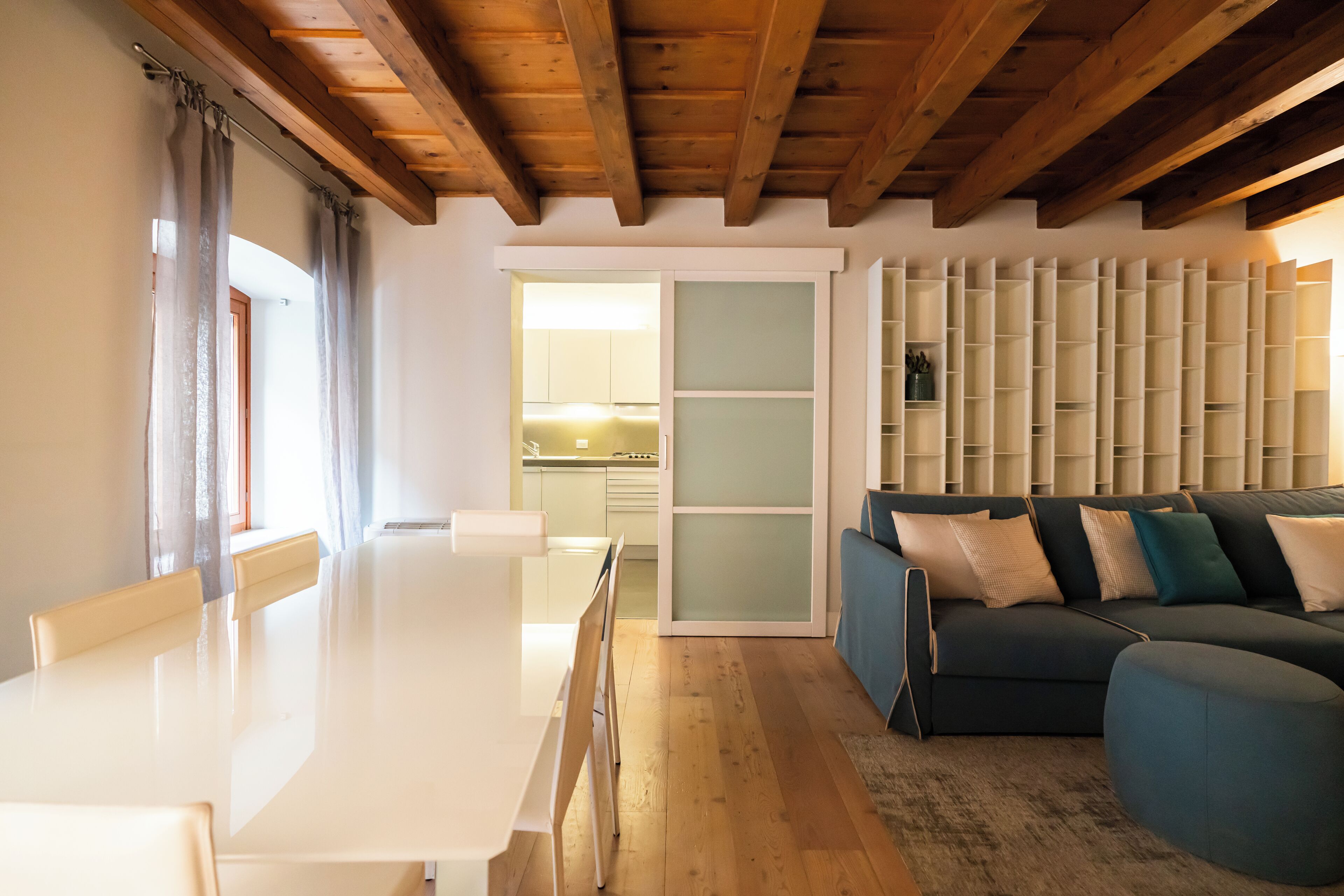 A modern living room with a wooden ceiling, a dining table with chairs, a sofa, and a built-in shelving unit.