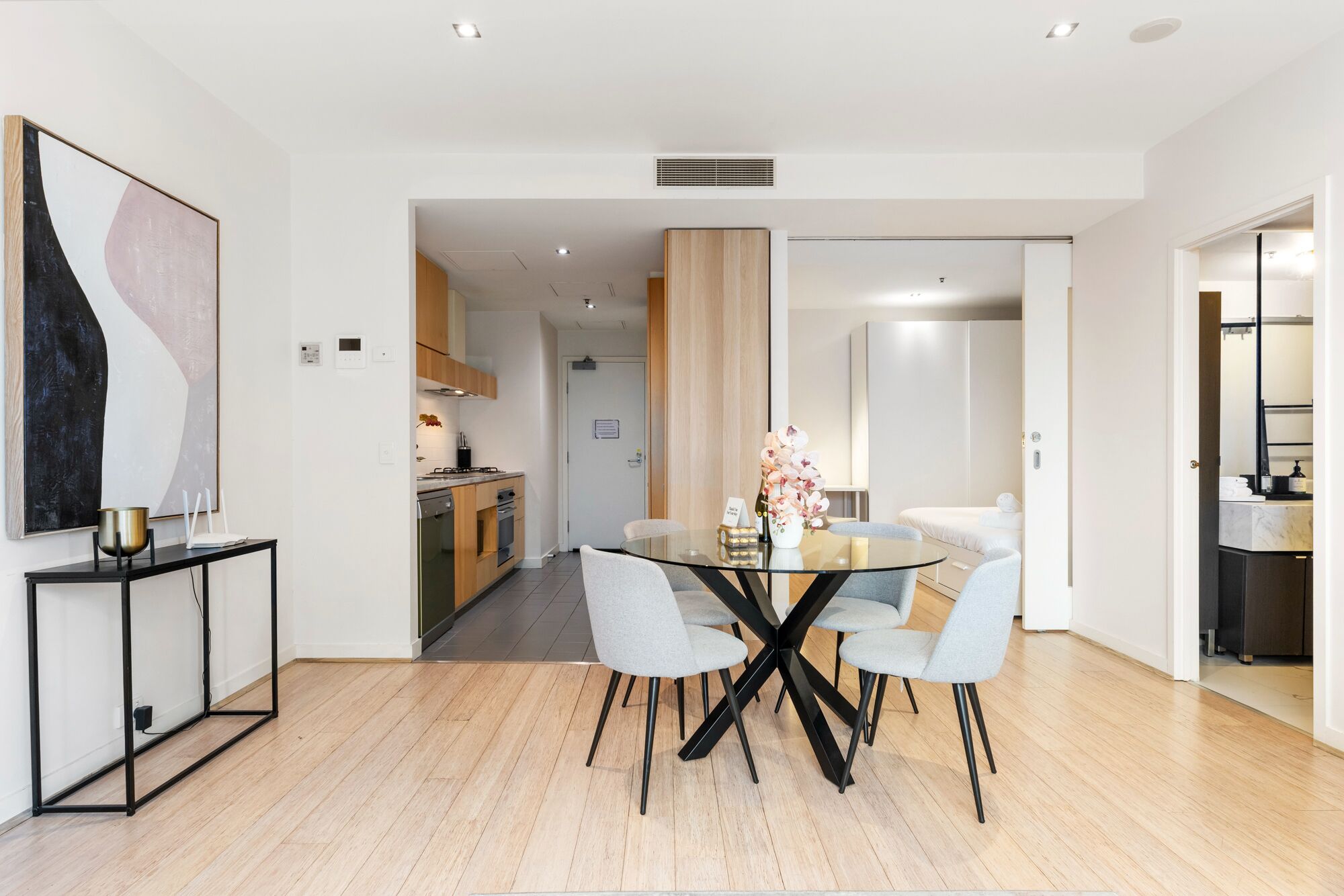 Un intérieur moderne avec un coin repas comprenant une table en verre et des chaises grises, une cuisine avec des meubles en bois et un couloir apparent menant à une autre pièce.