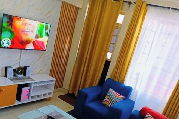 Ein Wohnzimmer mit an der Wand montiertem Fernseher, einem blauen Sessel, einer weißen Unterhaltungseinheit und einem Fenster mit Vorhängen.