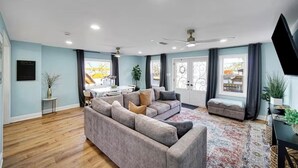 Una sala de estar moderna con un sofá seccional gris, un pequeño taburete gris, una alfombra estampada y una zona de comedor con mesa y sillas blancas.