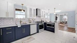 Una cocina moderna con armarios azul oscuro, encimeras blancas y una isla central. Hay luces colgantes, una ventana con vistas y un comedor visible al fondo.