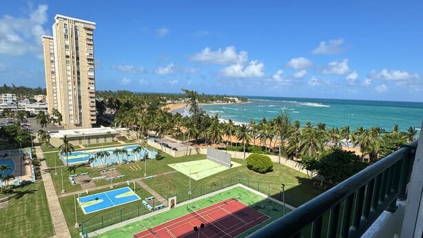 Vista desde un balcón que da a un complejo deportivo con canchas de tenis, piscina y playa.