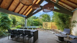 An outdoor patio with a wooden deck, a table with chairs, wicker hanging lights, and a view of a lush garden. - Villa Nature (Pougny)