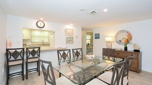 A dining area with a glass table and chairs, a kitchen in the background, and a decorative mirror on the wall. - Lovely, beachfront condo. Steps to Siesta Key #1 Beach in America. (Sarasota)