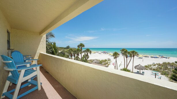 A balcony with blue chairs overlooking a beach with palm trees and umbrellas.