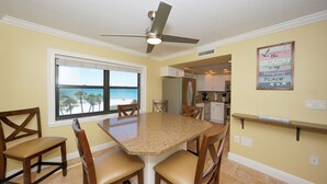A dining area with a large table and chairs, a ceiling fan, and a view of the beach through a window.