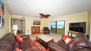 A living room with leather sofas, a coffee table, a recliner, a bookshelf, and a TV. There is a ceiling fan and a view of the beach through a sliding glass door.