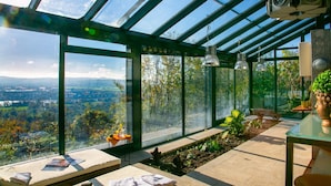 Une véranda dotée d’un toit en verre, offrant une vue sur un paysage montagneux. À l’intérieur, on trouve des coins salon avec des coussins et une petite table sur laquelle est posé un vase.