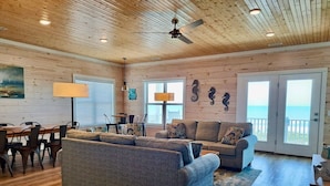 A spacious living room with a large sofa, a smaller sofa, and a dining area with chairs. There are two windows with a view of the ocean. - Stunning Oceanfront Retreat w/ Private Pool, Elevator, Handicap Accessibility (Ocean Isle Beach)