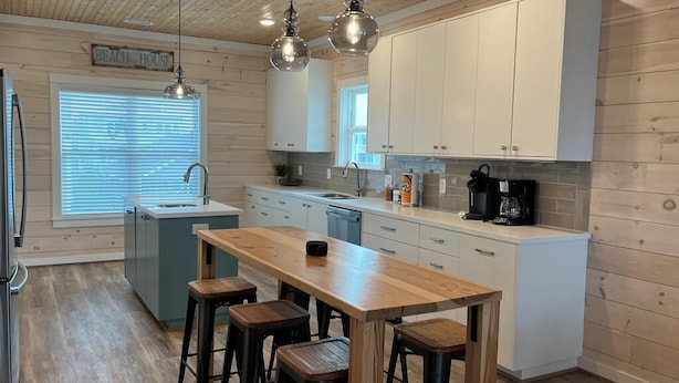 A modern kitchen with wooden flooring, white cabinets, a central island with stools, pendant lighting, and a window with blinds. - Stunning Oceanfront Retreat w/ Private Pool, Elevator, Handicap Accessibility (Ocean Isle Beach)