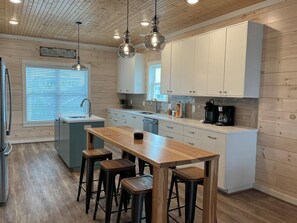 A modern kitchen with wooden flooring, white cabinets, a central island with stools, pendant lighting, and a window with blinds.
