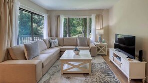 A living room with a beige sofa, a coffee table, a TV, and a balcony with curtains.