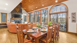 Un coin repas avec une table en bois dressée pour six personnes, entourée de chaises, avec une cheminée en pierre et un téléviseur en arrière-plan.