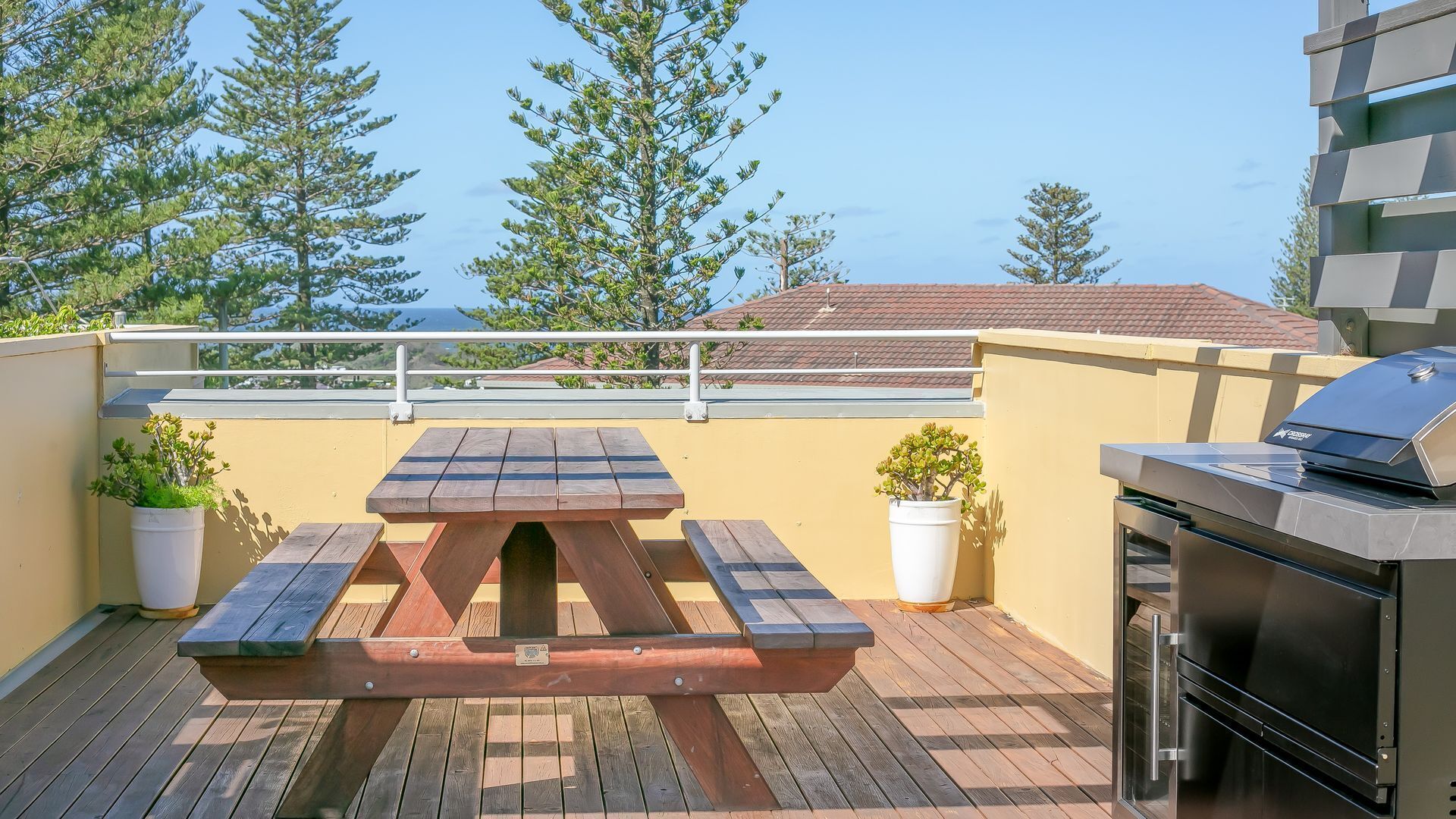 A rooftop patio with a wooden picnic table, potted plants, and a barbecue grill.