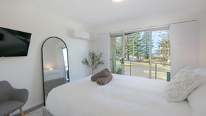 A modern hotel room with a large bed, a flat-screen TV, a grey armchair, and a balcony with a view of trees and a parking area. - Seaspray 6 - Short walk to the beach and Yamba's CBD (Yamba)