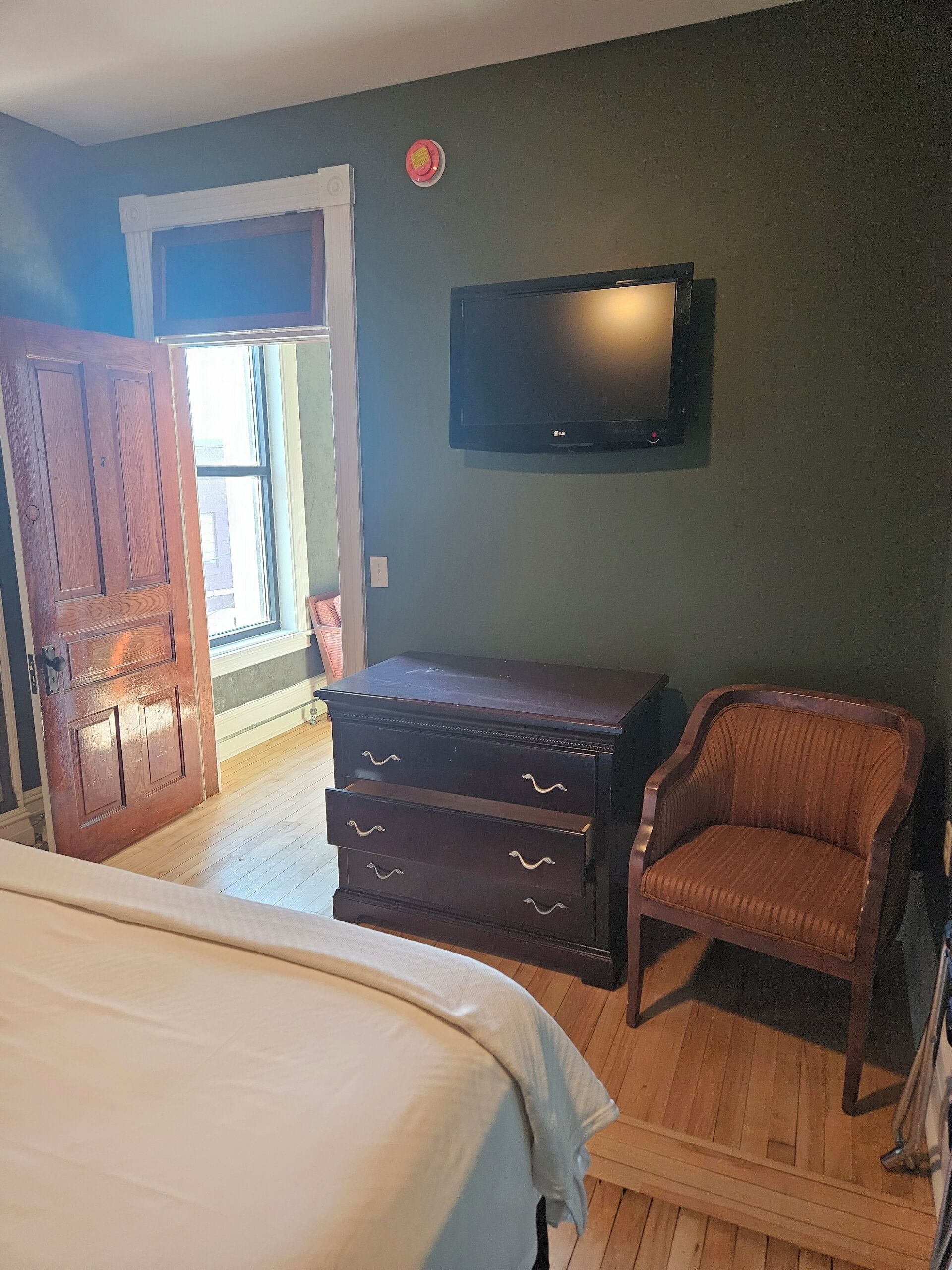 Une chambre à coucher comprenant un lit, une commode, une chaise, un téléviseur et une porte donnant sur une autre pièce.