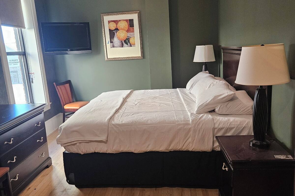 Un dormitorio con cama, dos lámparas de noche, ventilador de techo, televisor de pantalla plana, cómoda y un cuadro en la pared.