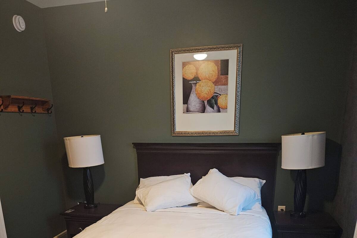 Una cama bien hecha en una habitación con pared verde, dos mesitas de noche con lámparas y un cuadro enmarcado sobre la cama.