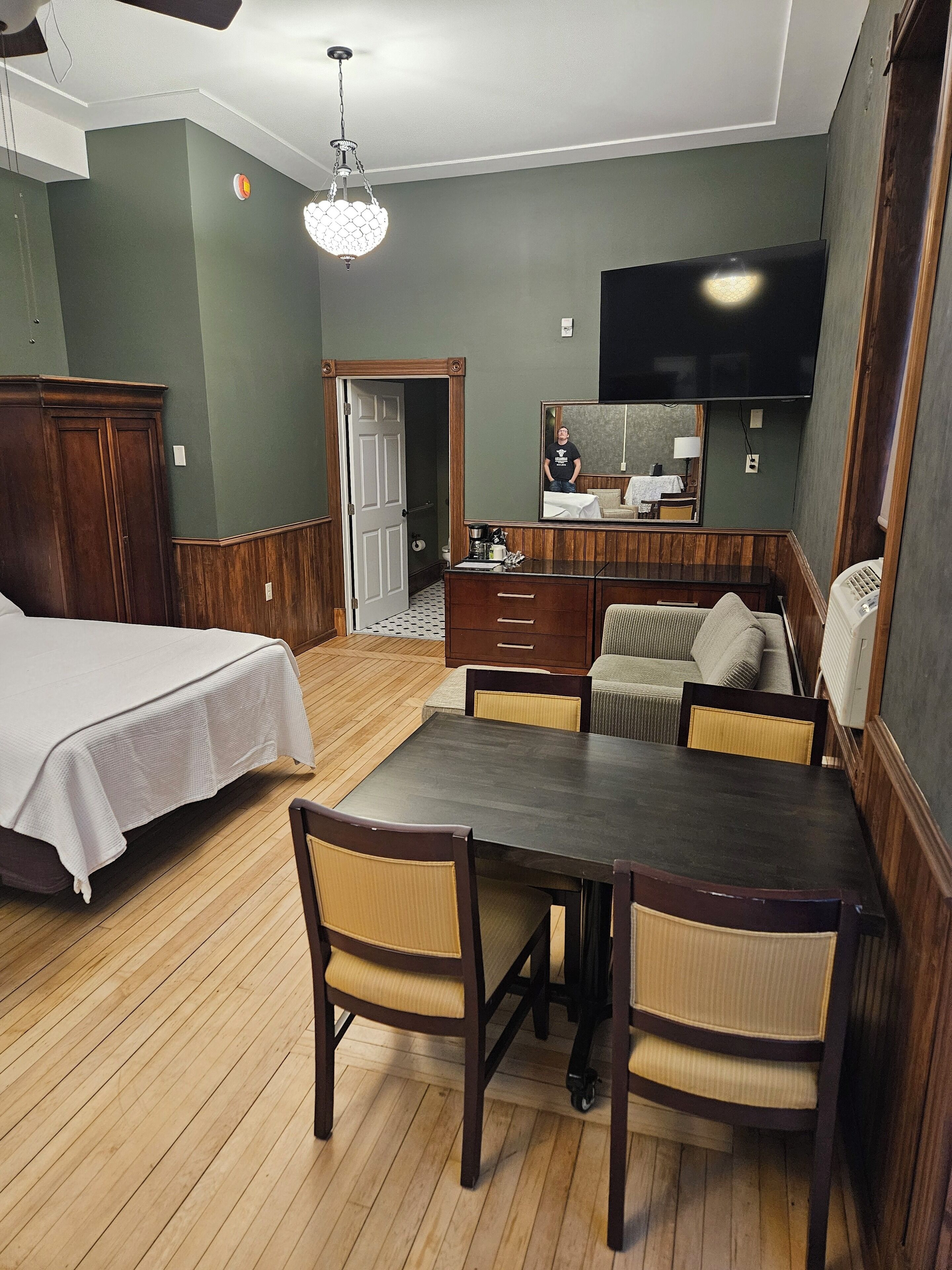 Una habitación de hotel con cama, una mesa de comedor de madera con sillas, un sofá, un televisor montado en la pared y un armario de madera.