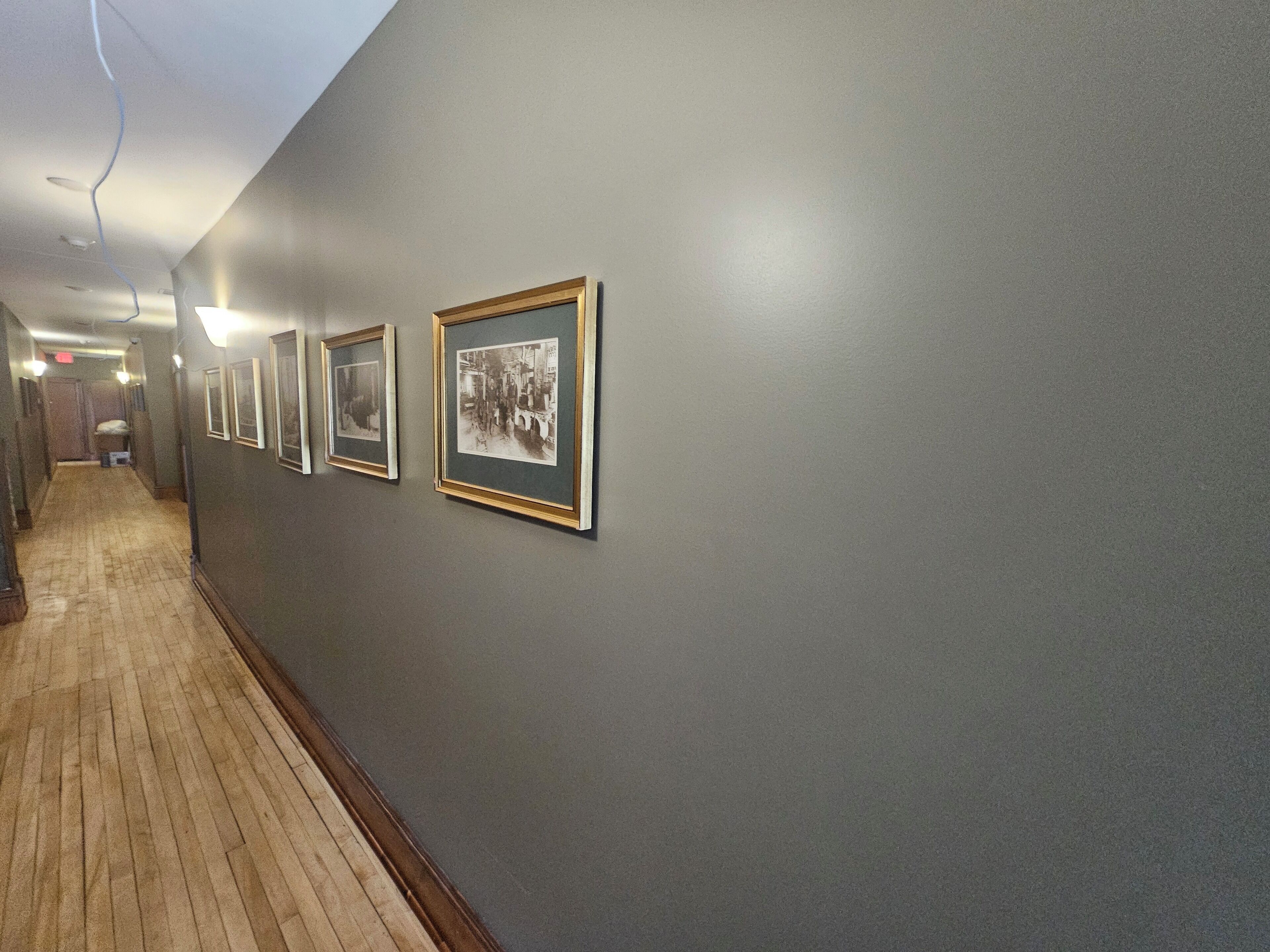 A hallway with framed pictures on the wall, wooden flooring, and a ceiling with exposed wiring.